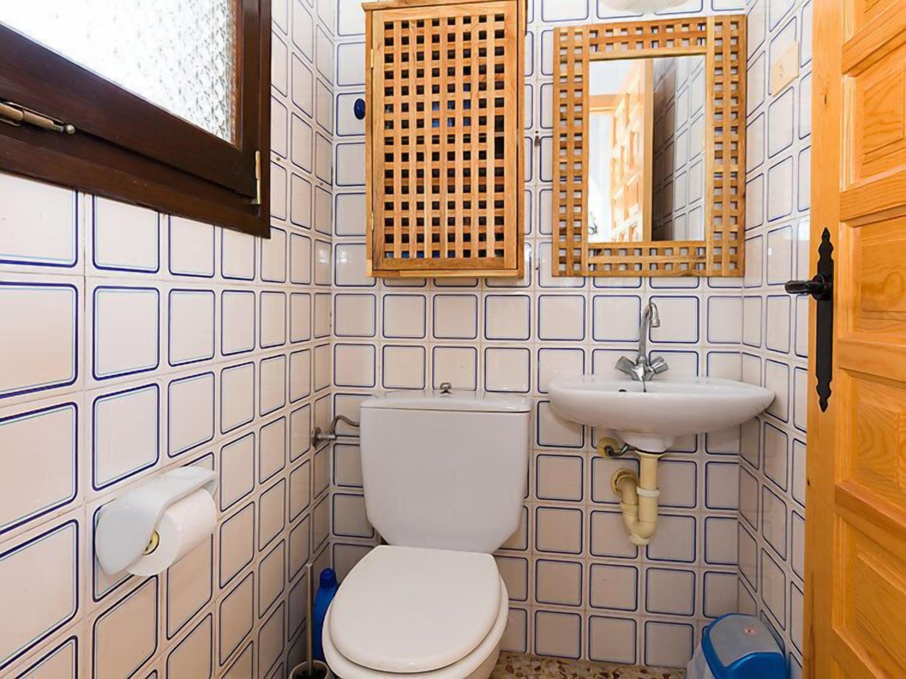 Kylpyhuoneen seinät ovat valkoisilla laatoilla, siinä on valkoinen wc-istuin, peilillä varustettu pesuallas ja puinen ovi.