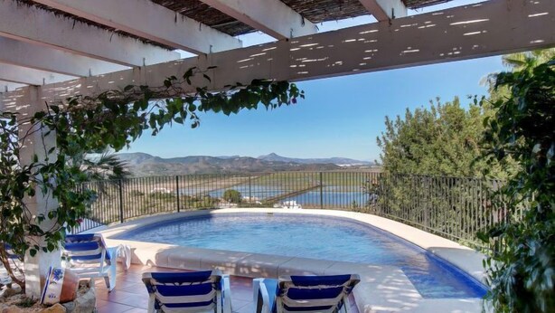 A swimming pool with two lounge chairs, surrounded by a fence and greenery, with a view of mountains and a body of water in the background.