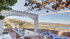 An outdoor patio with a table and chairs, a stone floor, and a view of a valley and river.