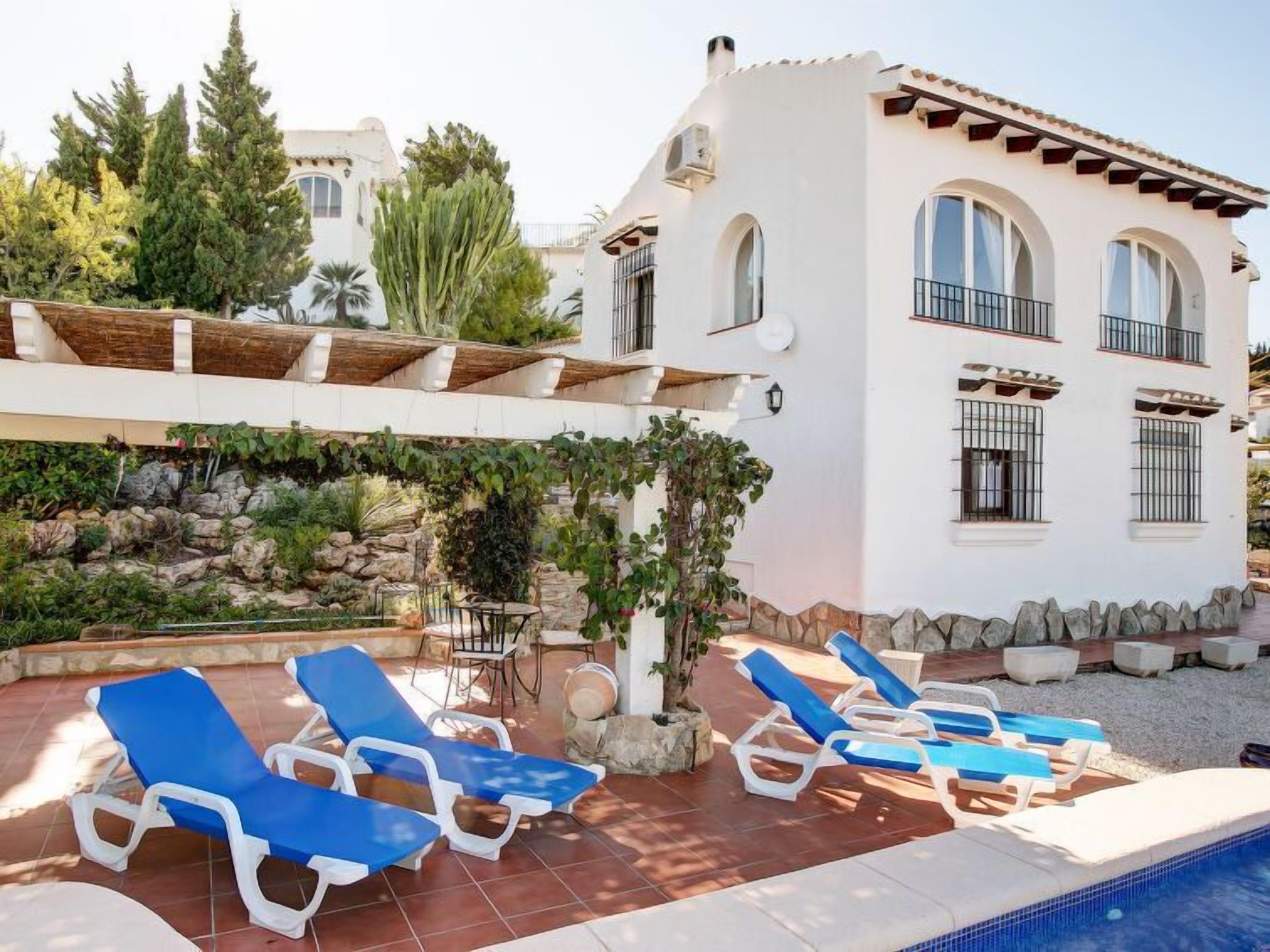 Una villa con piscina, sillas de descanso azules y un jardín con rocas y plantas.
