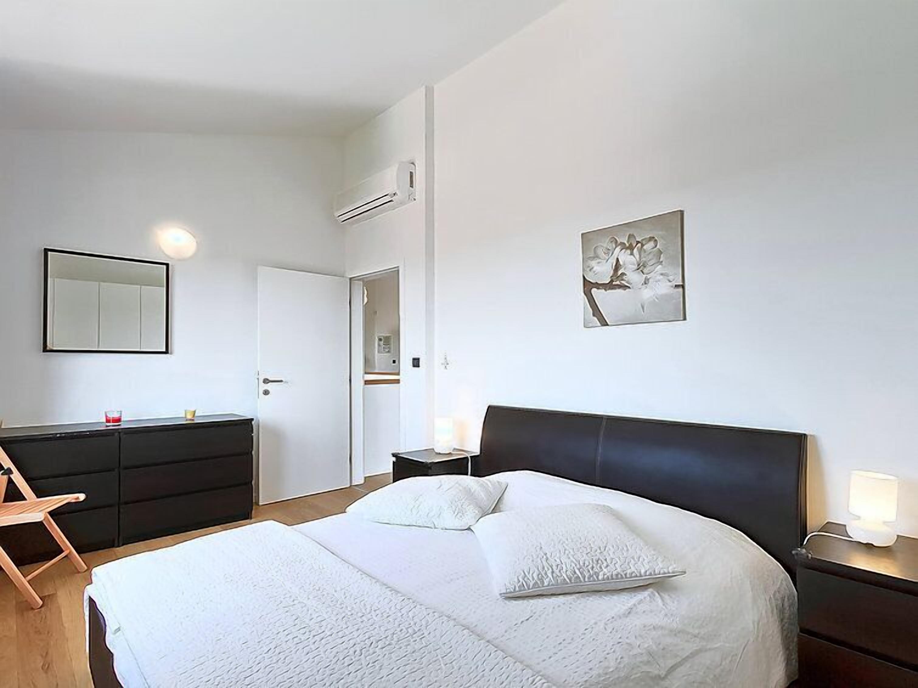 A bedroom with a bed, bedside tables, a dresser, a mirror, and a wall-mounted air conditioner.