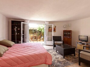 Ein Schlafzimmer mit einem Bett, einem Fernseher, einem Schreibtisch und einem Stuhl. Eine Schiebetür führt zu einem Balkon mit Tisch und Stühlen.