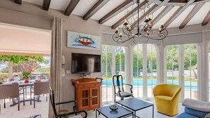 Ein geräumiger Wohnbereich mit einer großen Couch, einem gelben Sessel, einem Fernseher und einer Holzkommode. Es gibt einen Essbereich mit Korbstühlen und einem Tisch, sowie einen Blick auf einen Swimmingpool durch große Fenster.