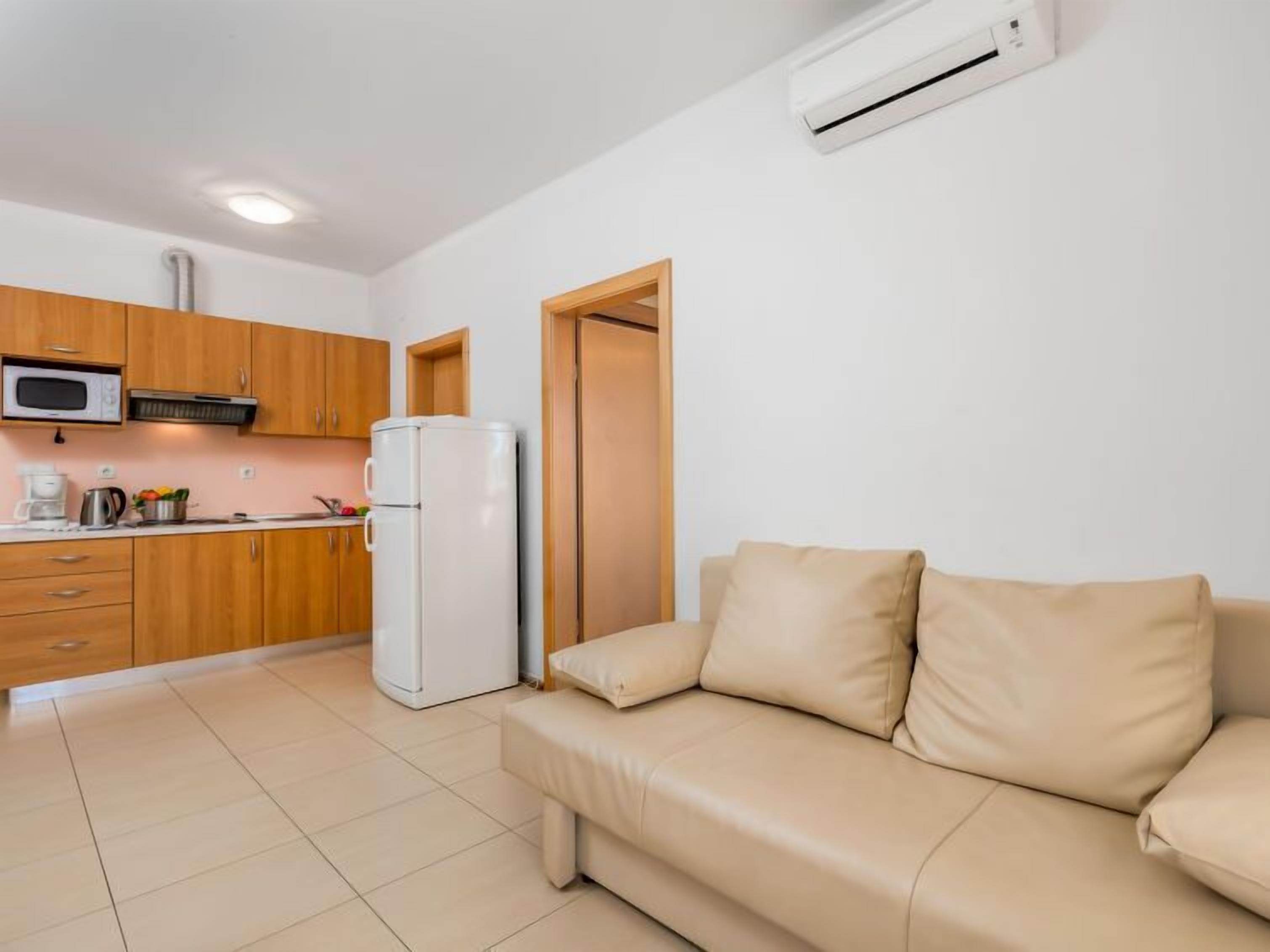 A compact living space with a kitchenette, a white refrigerator, a beige sofa, and a white air conditioning unit.