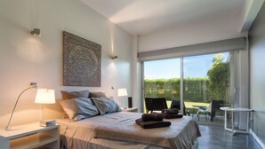 A modern bedroom with a large bed, two bedside lamps, a wall art piece, and a view of a garden through a sliding glass door. - Villa Vogue by Interhome (Ferragudo)