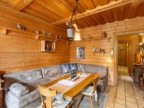 Ein gemütliches Blockhaus-Interieur mit Holzbalkendecke, einem großen Sofa, einem Esstisch mit Stühlen und einer kleinen Küchenzeile.