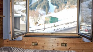 Une pièce dotée d’une grande fenêtre donnant sur un paysage enneigé, avec une rivière, des arbres et des bâtiments. On y trouve un lit recouvert d’une couverture à carreaux et un oreiller orné d’un motif en forme de cœur.