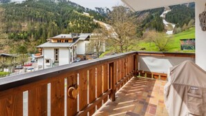 Un balcon doté d’une balustrade en bois, d’où l’on peut admirer un paysage montagneux parsemé de maisons et traversé par une route sinueuse.