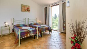 Un dormitorio con dos camas, un cuadro en la pared, una ventana con cortinas, un termostato y un jarrón con flores rojas.