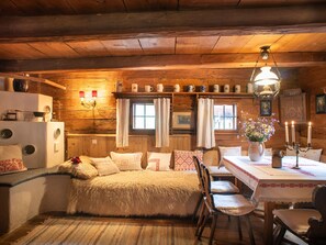 Ein gemütliches Blockhausszimmer mit einem Bett, einem Esstisch mit Stühlen, einer Holzbalkendecke und einem Fenster mit Vorhängen.