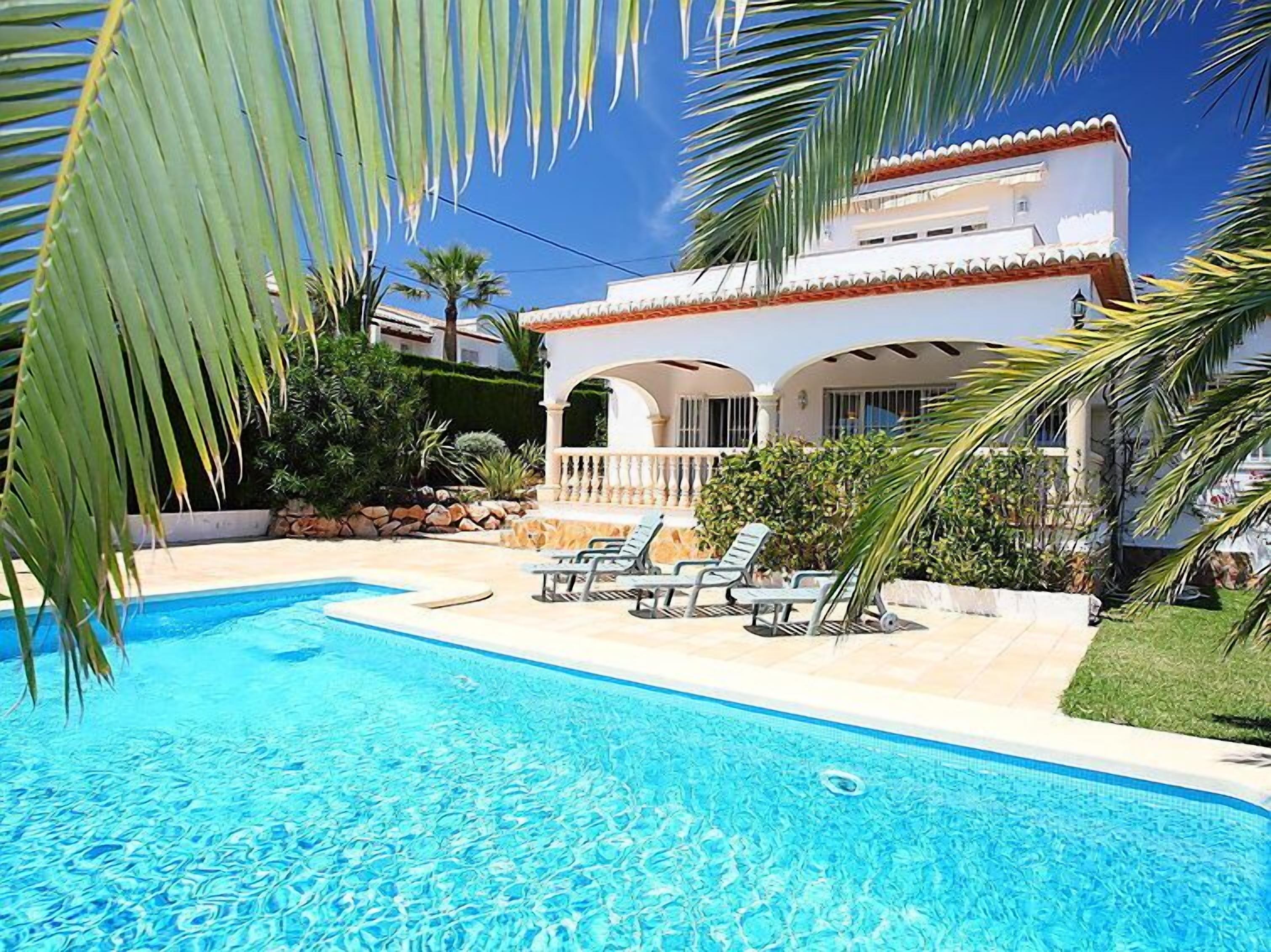 A pool area with lounge chairs, a white building with arches, and palm trees.