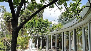 A covered patio with glass walls, surrounded by lush greenery and a large tree. - Vacation home Agora by Interhome (Nice)