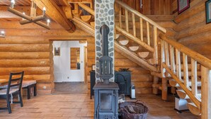Interno di una baita di legno con camino centrale, una scala in legno e un'area pranzo con una sedia e un tavolo.