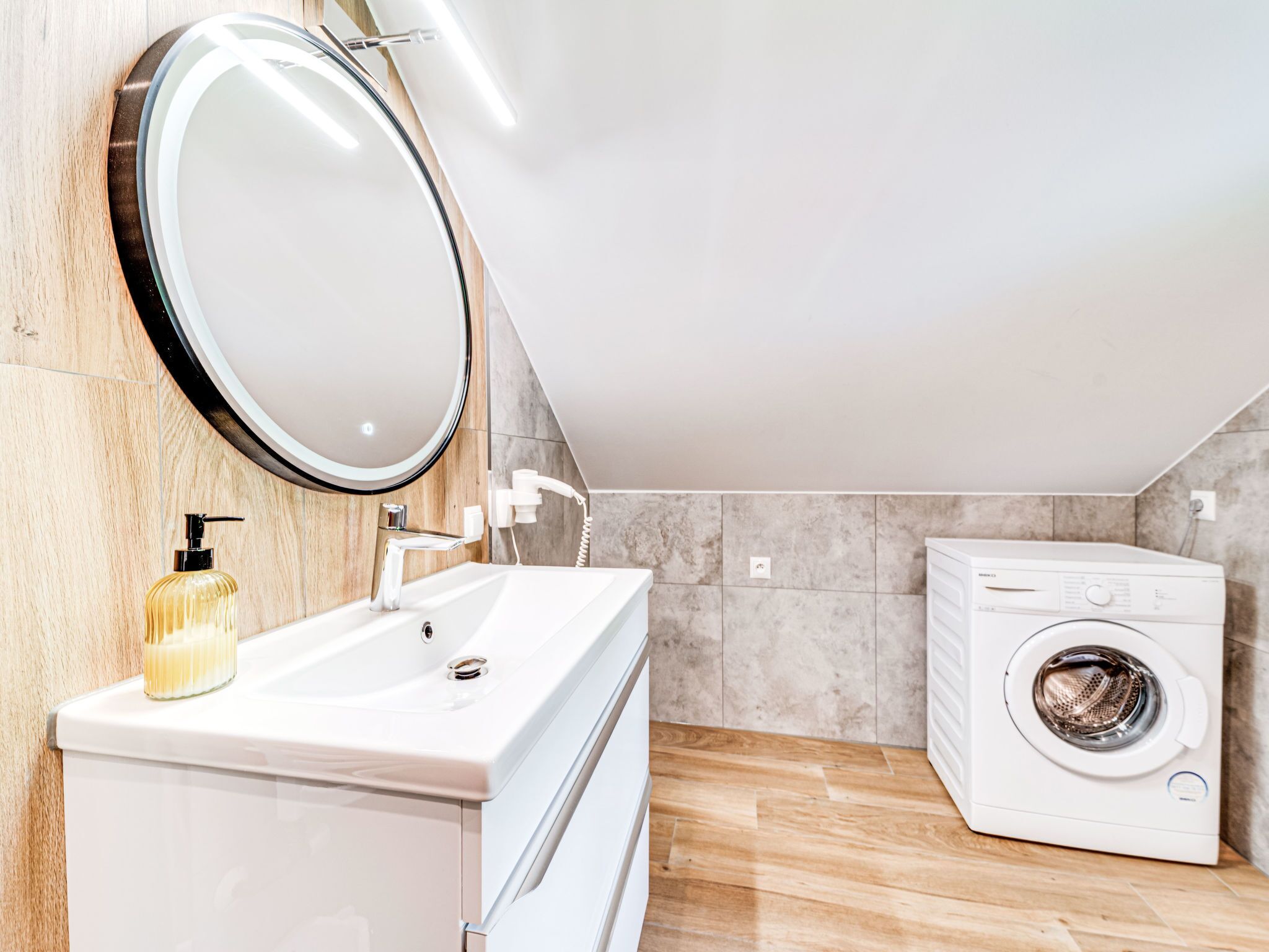 A modern bathroom with a white sink, a round mirror, a handheld showerhead, and a front-loading washing machine.