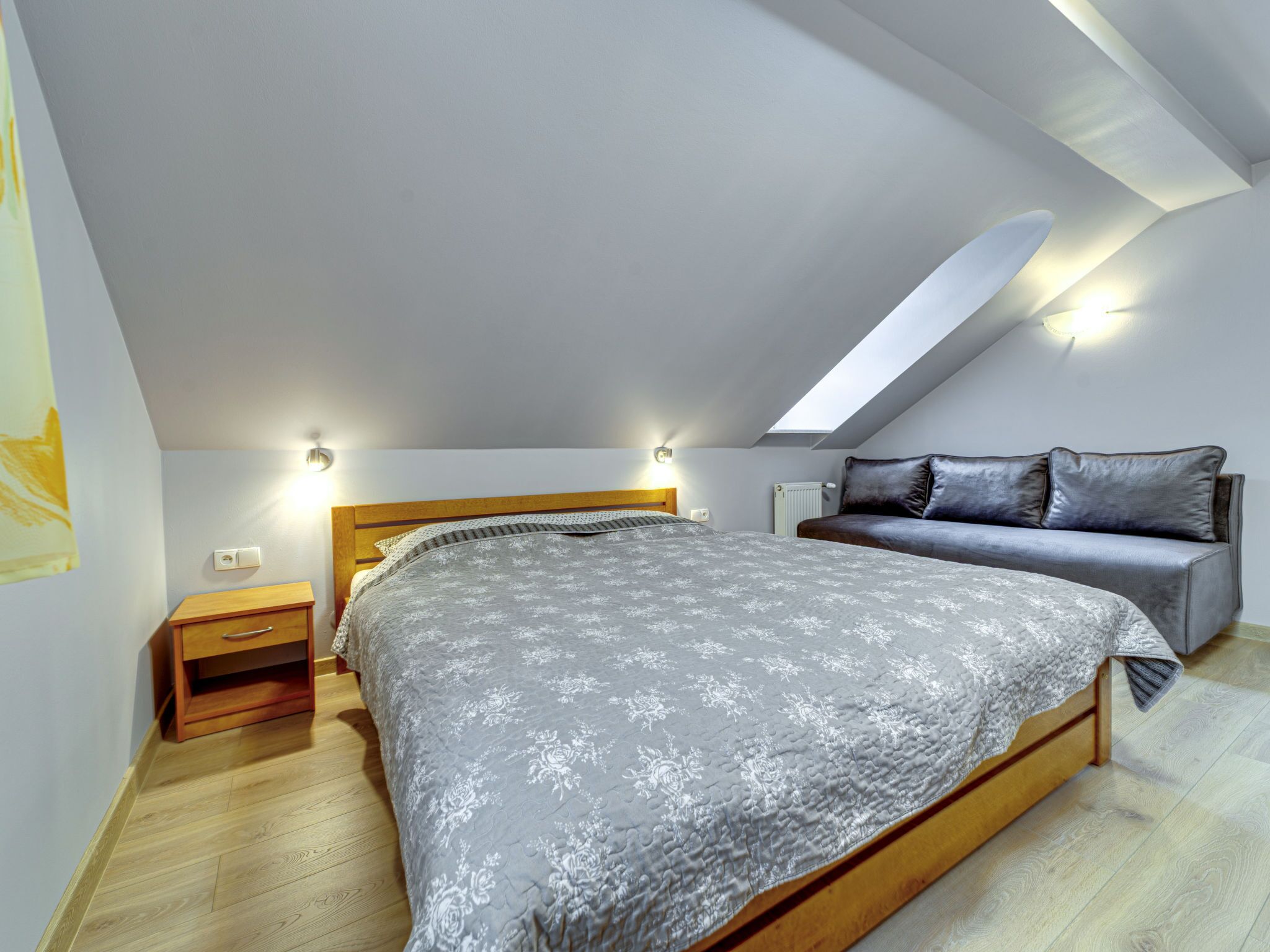 A bedroom with a sloped ceiling, a bed with a grey floral bedspread, a wooden nightstand, a grey sofa, and a skylight.