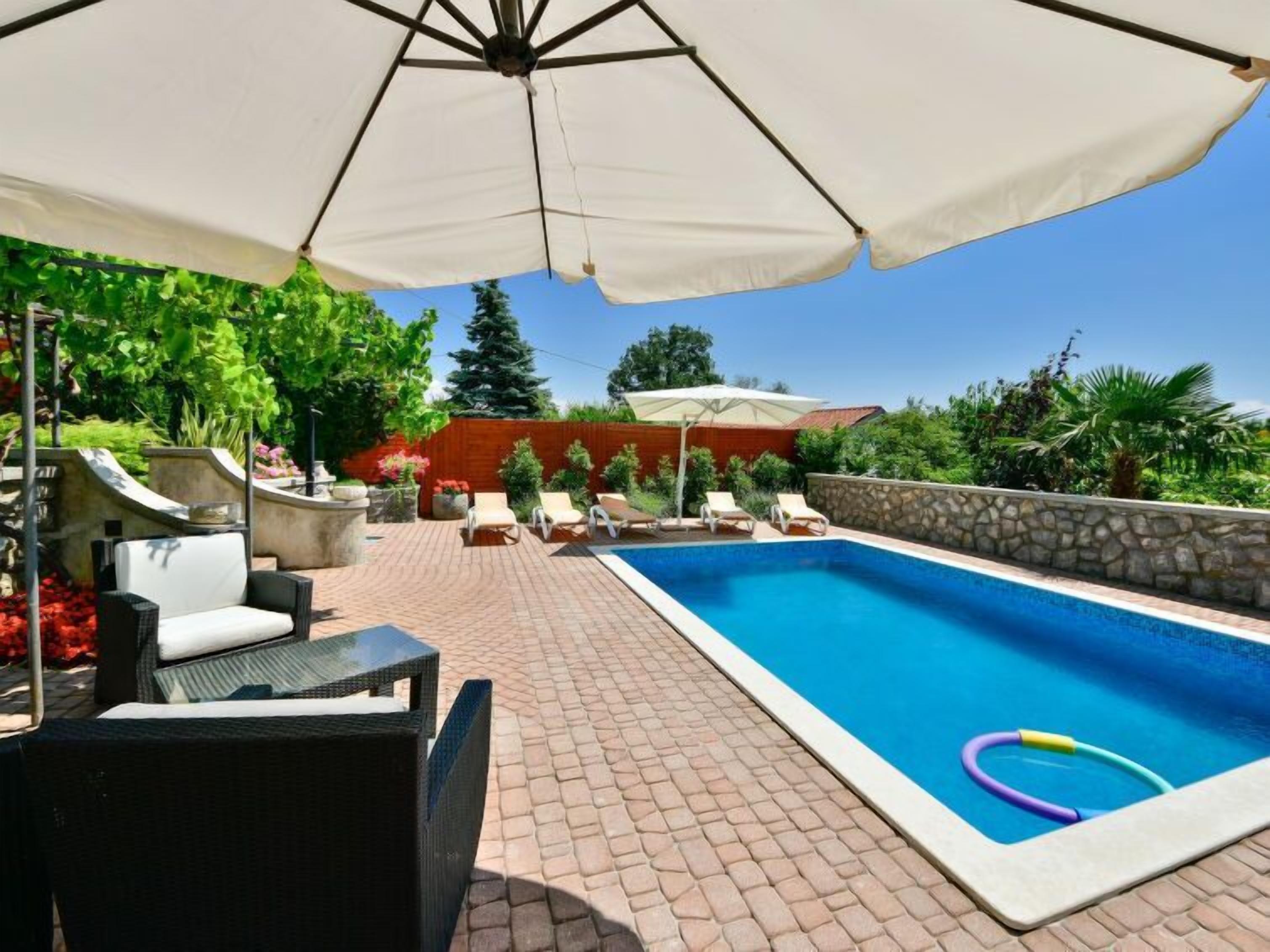 Une piscine entourée de verdure et d’un mur de pierre, avec des chaises longues et un parasol.