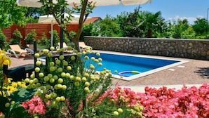 Une piscine entourée d’une végétation luxuriante et de fleurs, avec un mur de pierre et des chaises longues en arrière-plan.