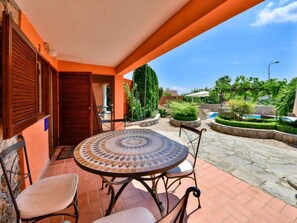 Une terrasse avec une table et des chaises carrelées, une porte en bois et une vue sur un jardin aménagé avec une piscine.