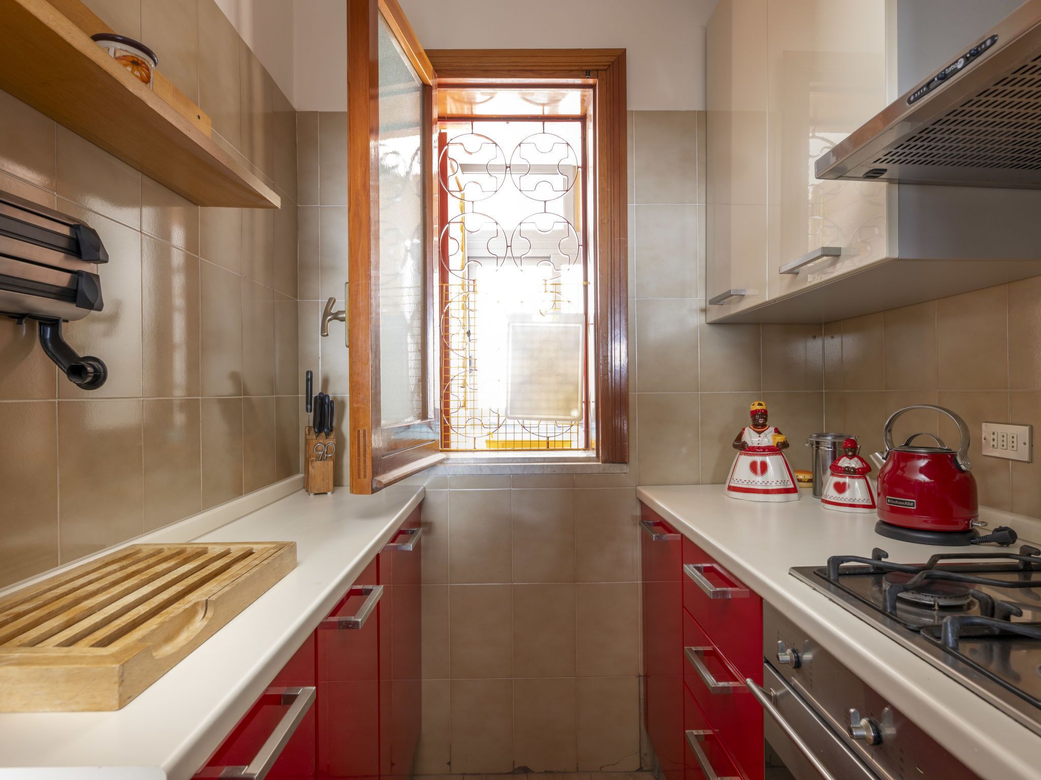 Une cuisine équipée d’une cuisinière à gaz, de placards rouges, d’une planche à découper en bois et d’une fenêtre ornée d’un paravent métallique décoratif.