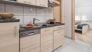 Een moderne keuken met een inbouwvaatwasser, een magnetron en een kookplaat. Er is een raam met gordijnen, en een bed is zichtbaar in de aangrenzende kamer.