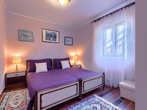 Ein Schlafzimmer mit einem Bett, zwei Nachttischlampen, einem Nachttisch, einem Fenster mit leichtem Vorhang und einem gemusterten Teppich.