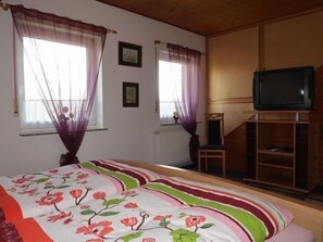 A bedroom with a bed, a television on a wooden stand, and two windows with sheer curtains.