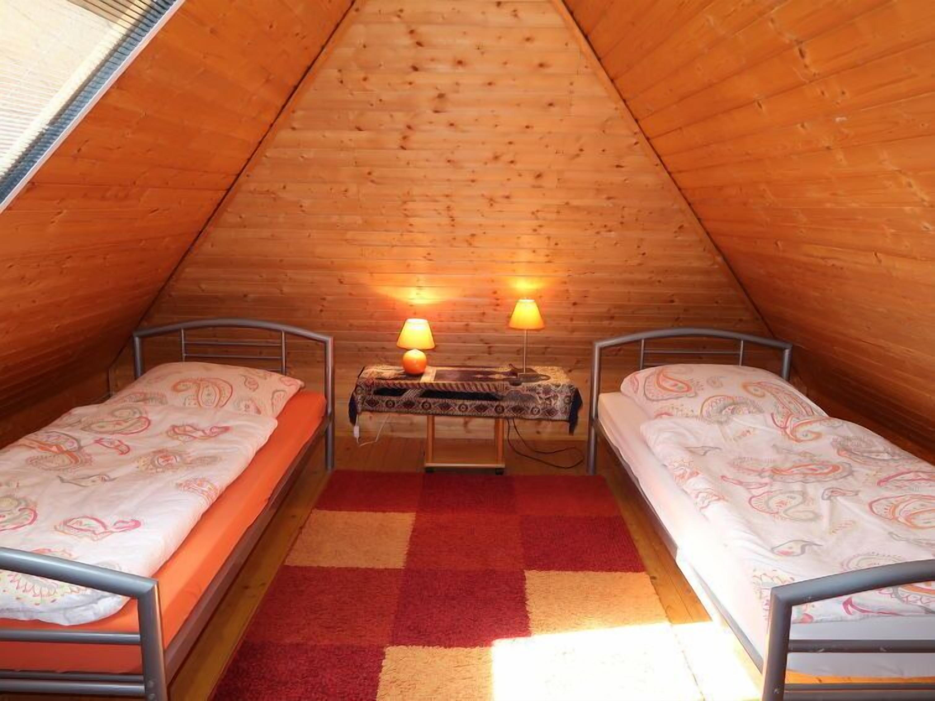 Un pequeño y acogedor dormitorio en el ático con dos camas individuales, una alfombra de cuadros rojos y beige, y dos lámparas de noche.