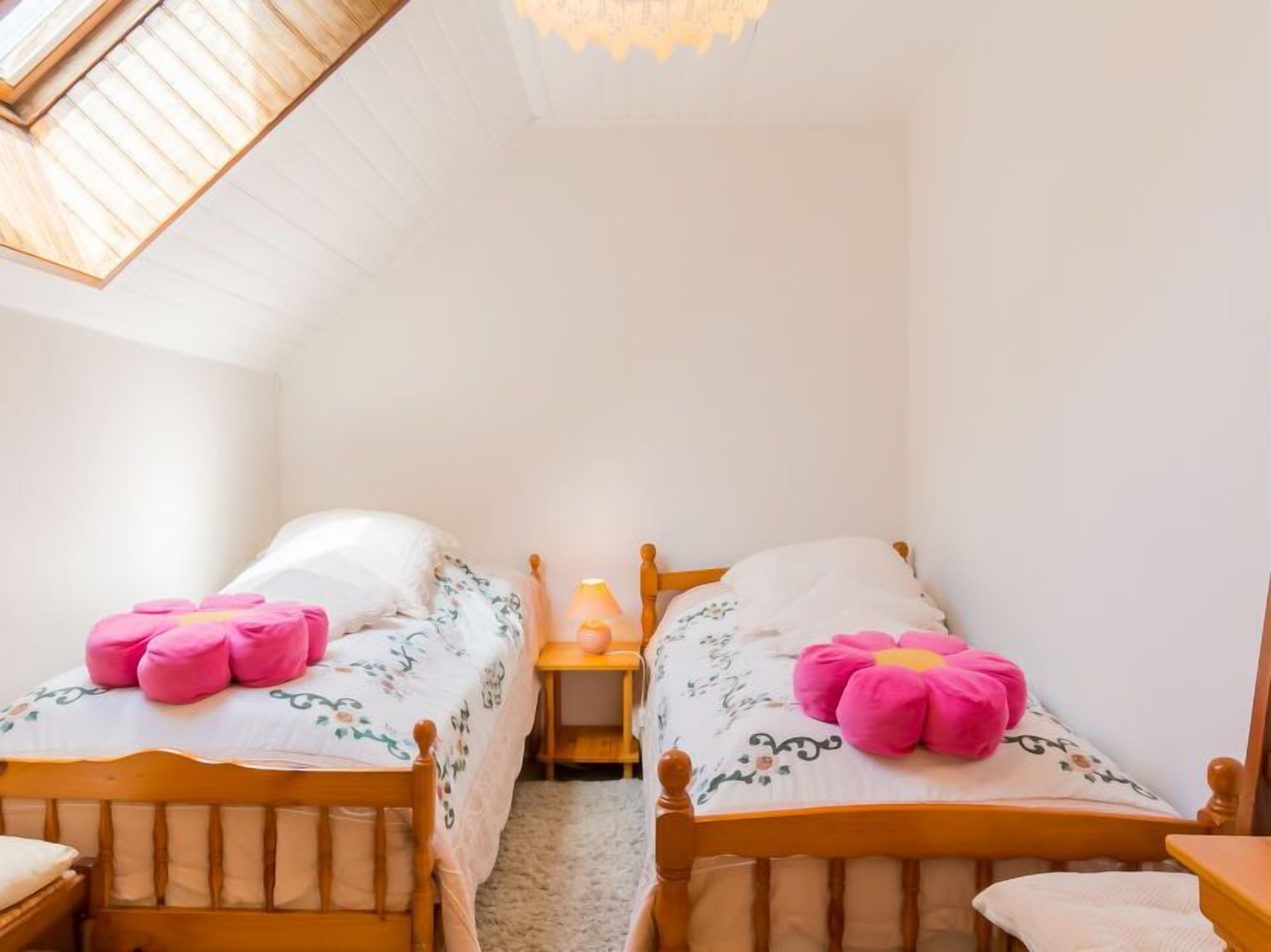 Um quarto pequeno e acolhedor com duas camas de madeira, cada uma com roupa de cama floral e almofadas cor-de-rosa. Uma pequena mesa de cabeceira com um abajur fica entre as camas.