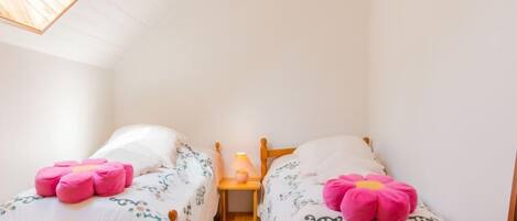 Um quarto pequeno e acolhedor com duas camas de madeira, cada uma com roupa de cama floral e almofadas cor-de-rosa. Uma pequena mesa de cabeceira com um abajur fica entre as camas.