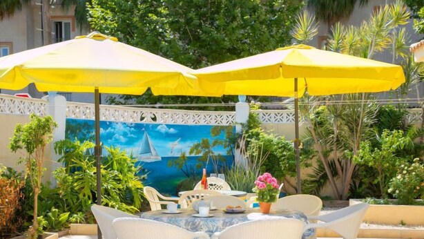 An outdoor dining area with white wicker chairs and yellow umbrellas, surrounded by lush greenery and a mural depicting a sailboat on the sea. - Vacation home Pedro by Interhome (Albir)