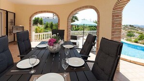 Un área de comedor exterior con una mesa preparada para dos personas, rodeada de sillas, un jarrón decorativo con flores y vistas a una piscina y al paisaje.