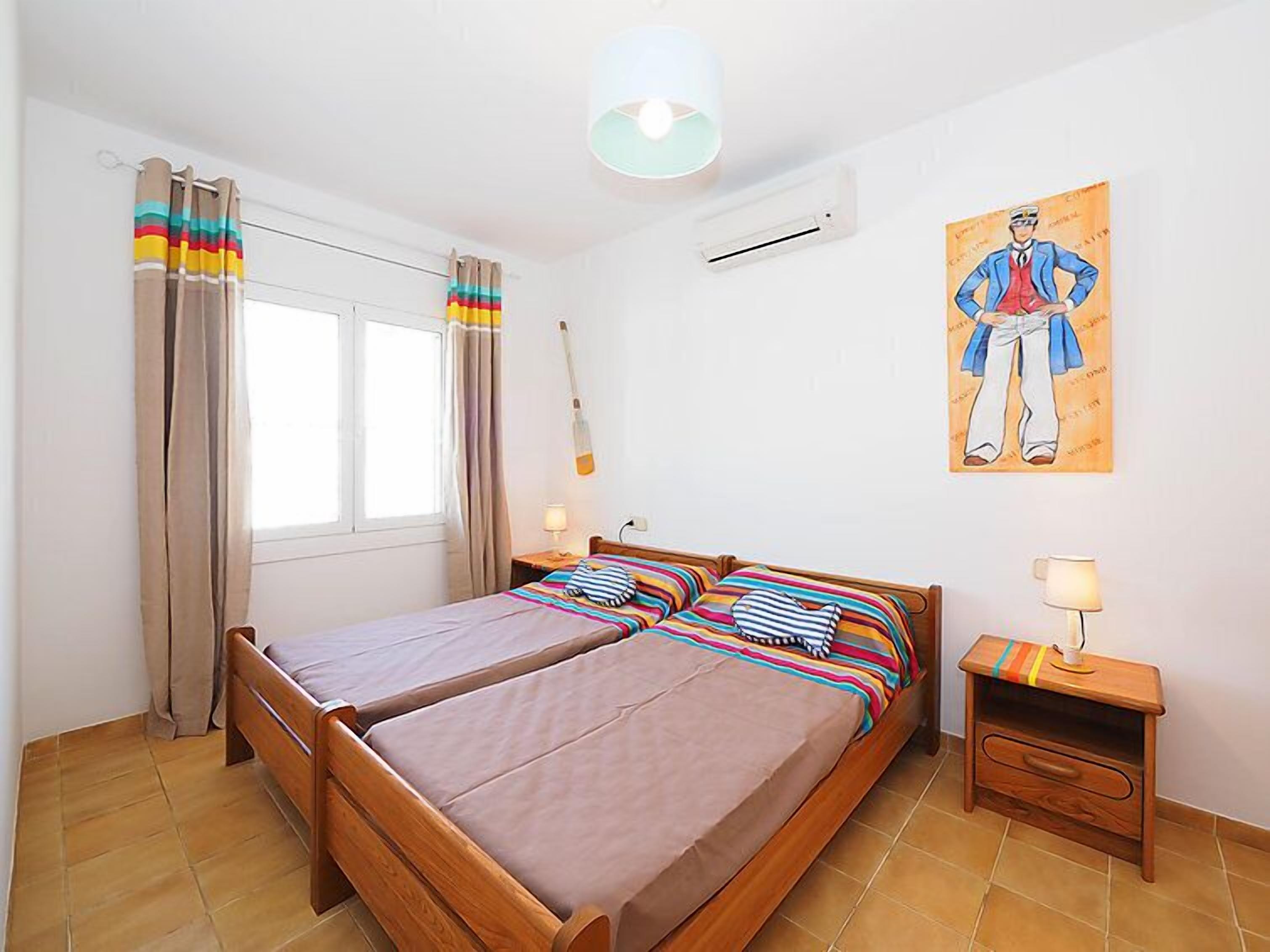 Una cama doble con ropa de cama a rayas, una mesita de noche con lámpara, aire acondicionado instalado en la pared y una ventana con cortinas.