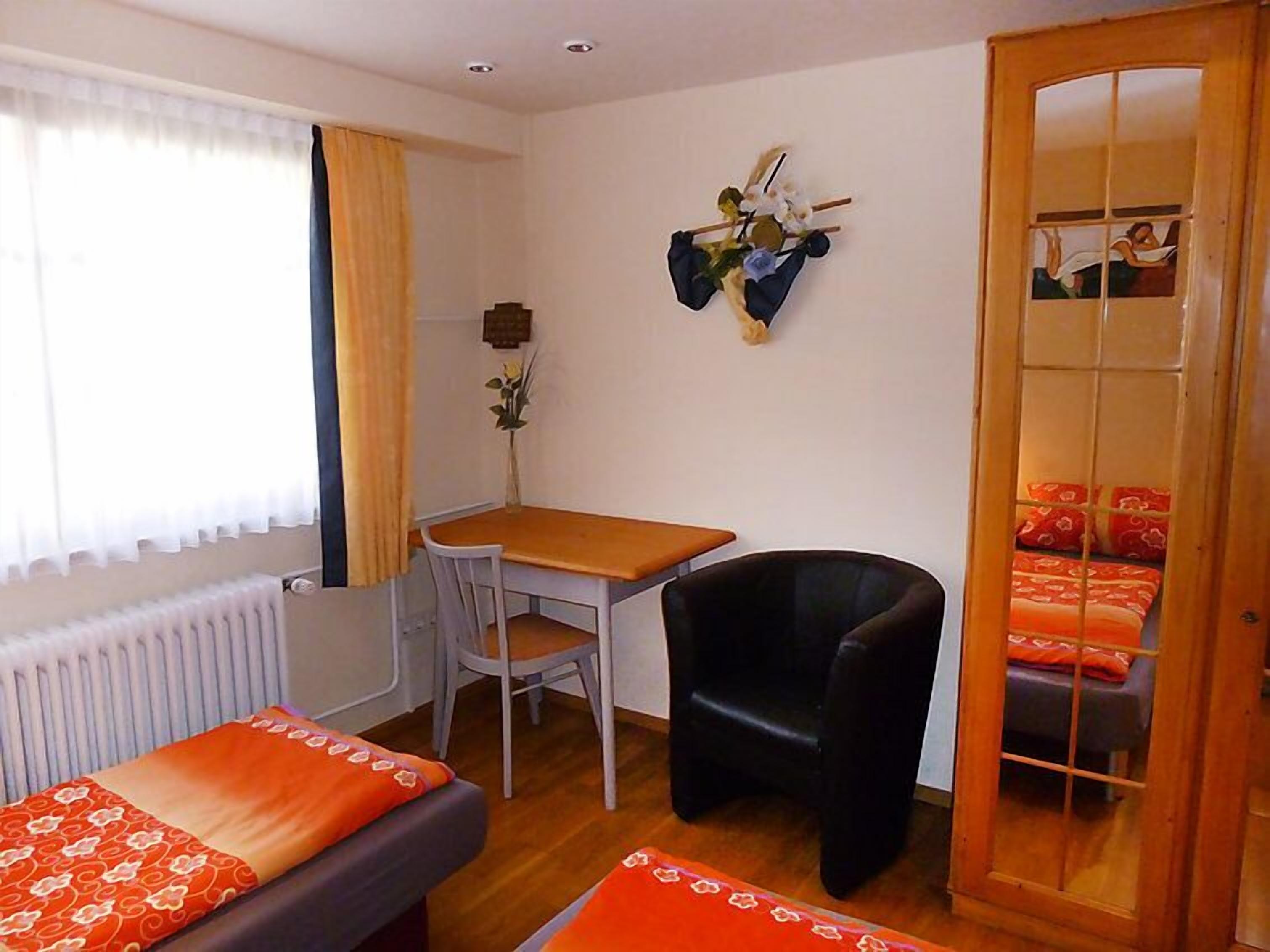 Una habitación con una mesa de madera, una silla negra, una cama con ropa de cama naranja y un espejo en la pared.