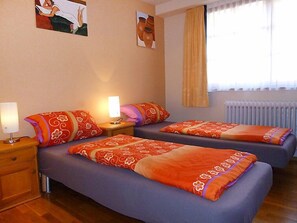 A hotel room with two beds, a nightstand with a lamp, a radiator, and a window with curtains.