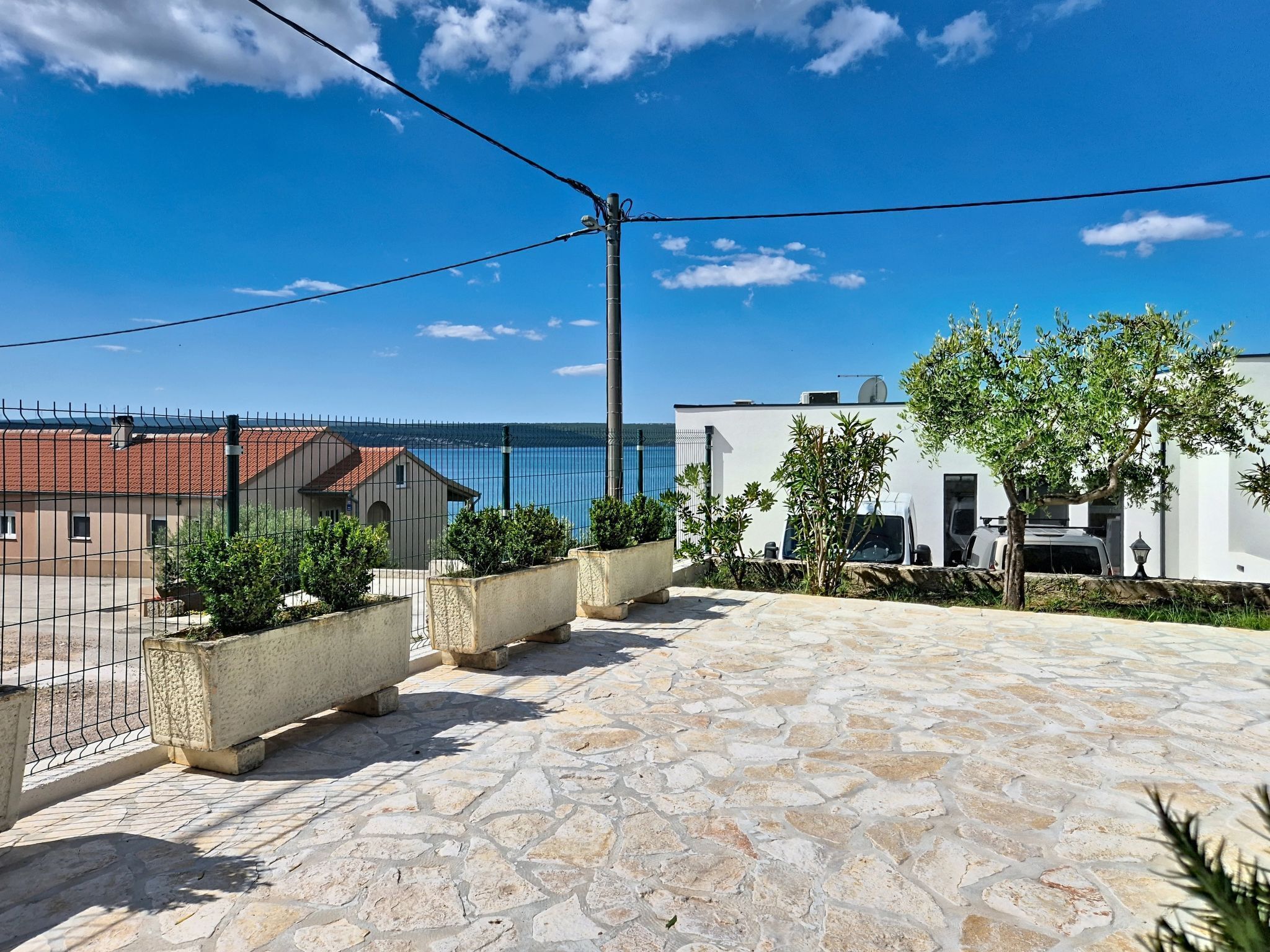 A rooftop terrace with stone flooring, a metal fence, and a view of the sea and buildings.