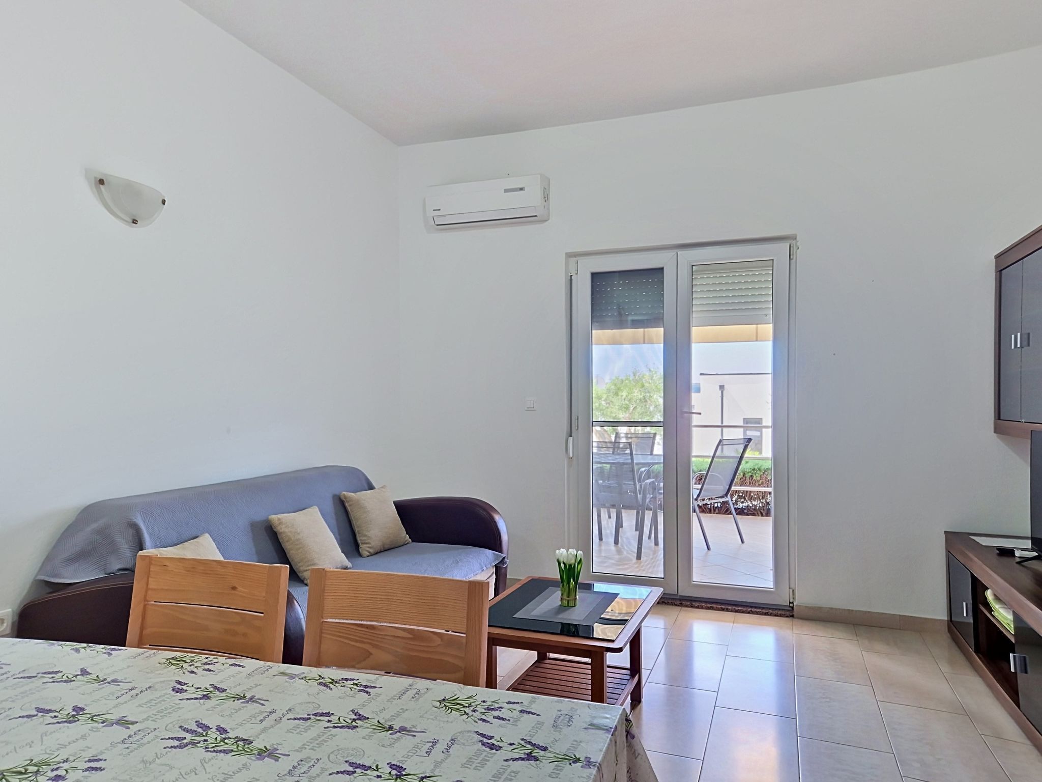 Una habitación con cama, sofá, una mesa con un florero, televisor y una puerta corrediza de vidrio que da a un espacio exterior.