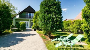 A two-story house with a paved driveway leading to a front door. There are two green lawn chairs on the grass to the right.