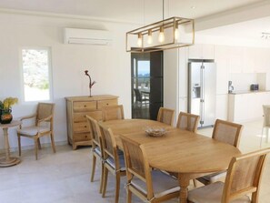 A modern dining area with a wooden table and chairs, a decorative light fixture above, and a view of the kitchen and dining room through an open door.
