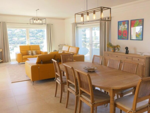A spacious living area with a yellow sofa set, a wooden dining table with chairs, and a large window offering a view of the outdoors.