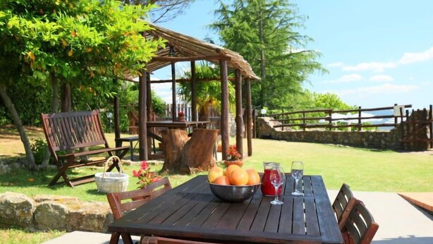 A wooden table with chairs, a basket of fruit, a pitcher, and a glass of red drink, set outdoors with a wooden pergola and a stone wall in the background.