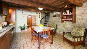 A rustic kitchen with a stone wall, wooden ceiling beams, a dining table with chairs, and a wooden bench.