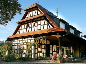 Une maison traditionnelle à colombages, dotée d’un toit à forte pente, d’une cheminée bien visible et d’un porche à l’avant, avec des chaises et une table.