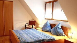 A bedroom with a sloped ceiling, a bed with blue bedding, two pillows, a chair, a nightstand, and a window with curtains. - Dieboldshof by Interhome (Ichenheim)