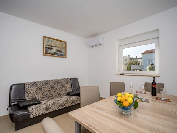 Ein modernes Wohnzimmer mit einer Couch, einem Esstisch mit Obstschale und Weingläsern, einem Bild an der Wand und einem Fenster mit Blick auf Gebäude.