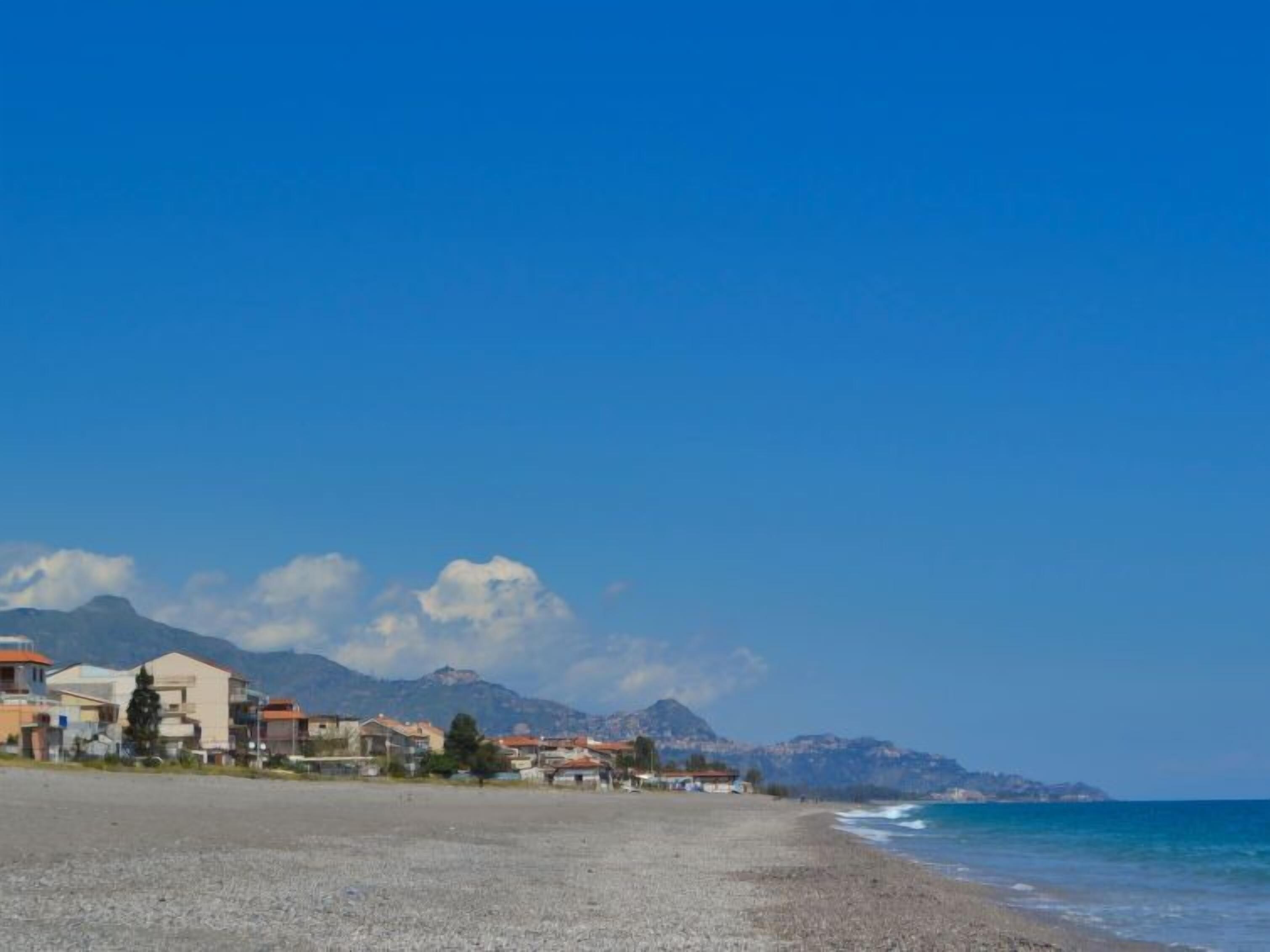 Una zona costiera con edifici lungo la riva, cielo sereno e montagne sullo sfondo.