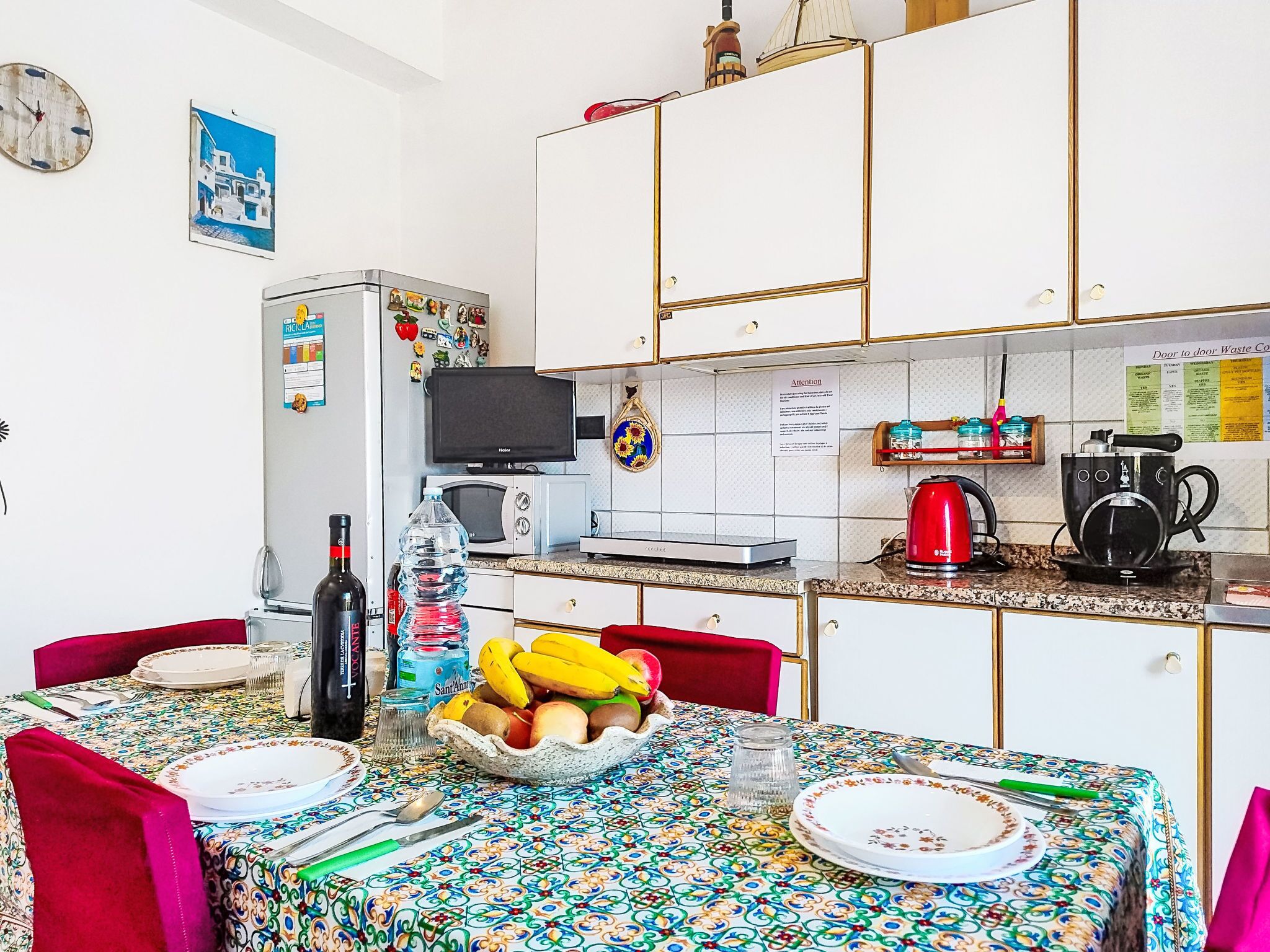 A compact kitchen with white cabinets, a microwave, a toaster, and a kettle. There is a dining table with a colorful tablecloth and a bowl of fruit.