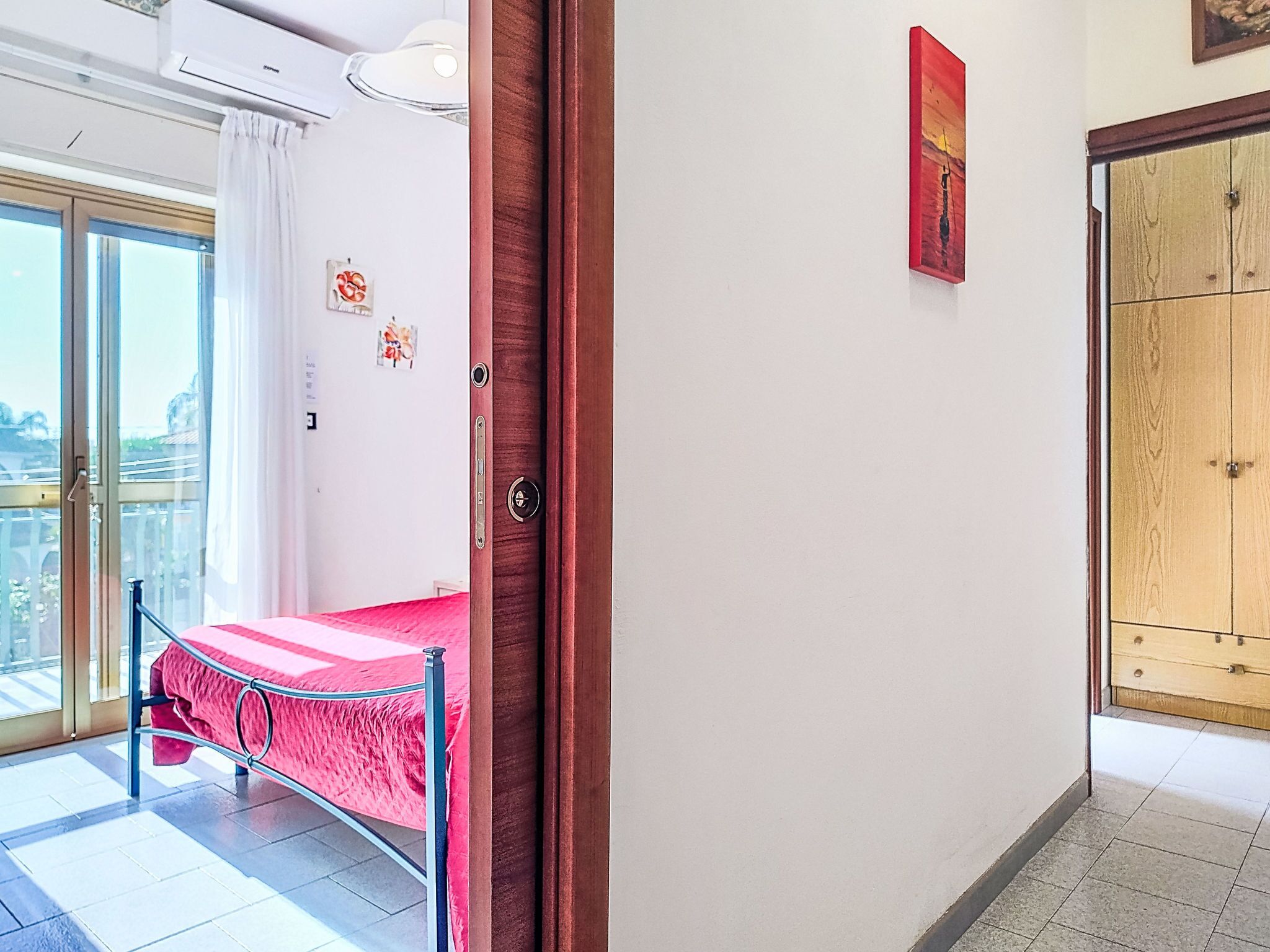 A room with a bed, a sliding glass door to a balcony, a wooden wardrobe, and a wall-mounted artwork.