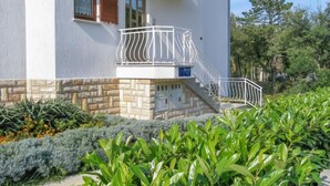 Un edificio de dos pisos con balcón, base de piedra y un jardín con diversas plantas.