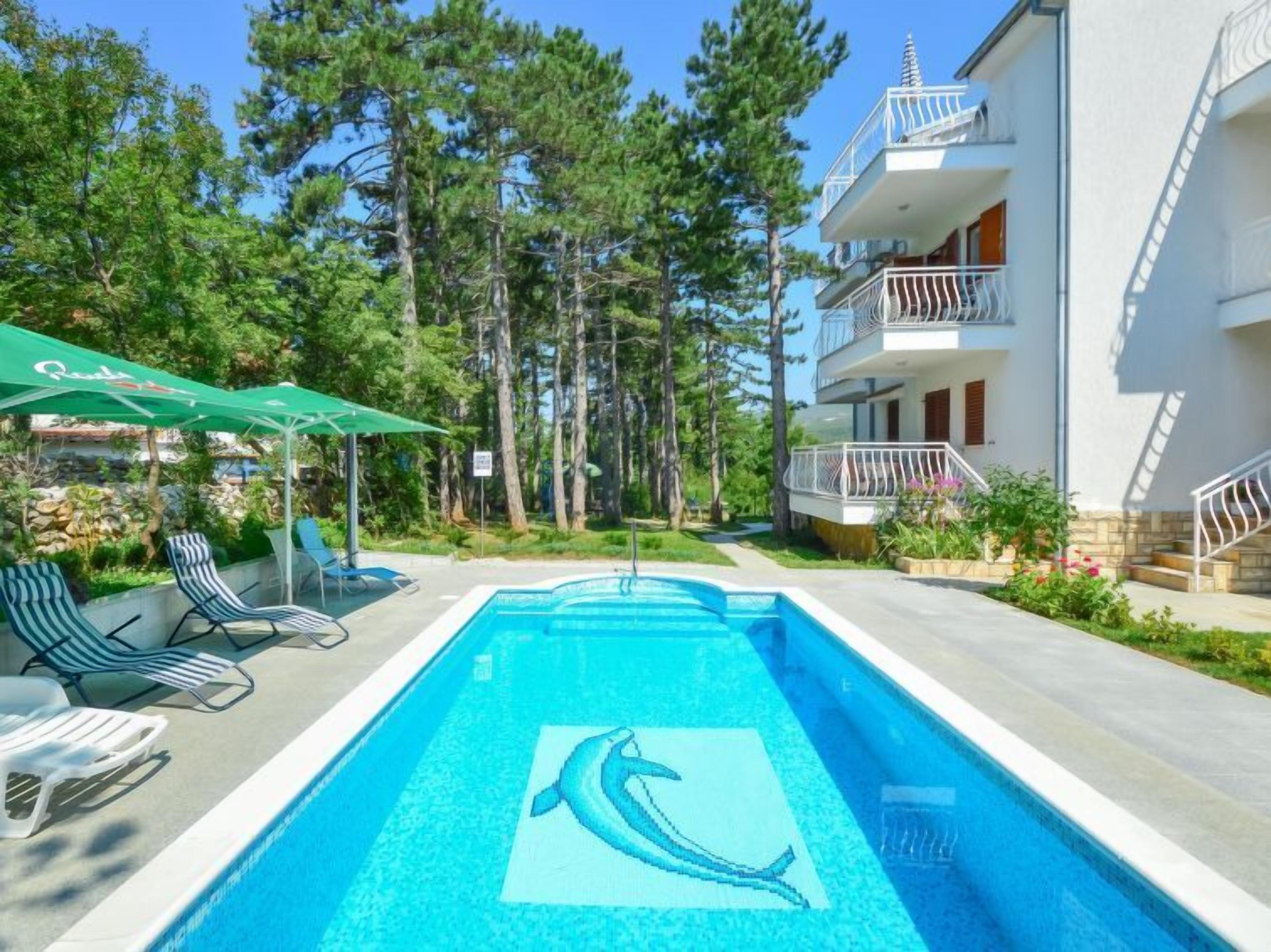 A swimming pool with a dolphin design, surrounded by lounge chairs and umbrellas, with a multi-story building in the background.