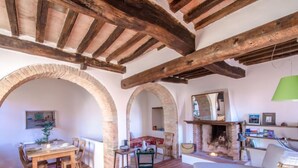 Ein rustikales Interieur mit freiliegenden Holzbalken, einem Essbereich mit Tisch und Stühlen, einem weißen Sofa und einem Kamin.